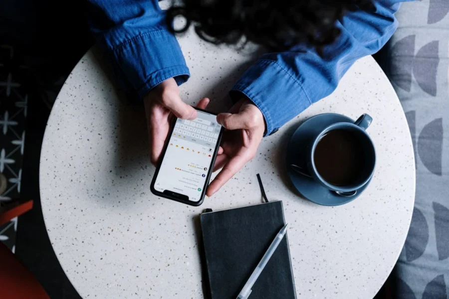 Persona escribiendo en su teléfono móvil, sentada a una mesa con una taza de café y una libreta.