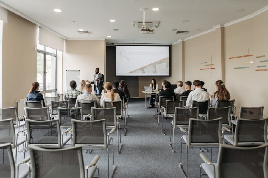 Conferencia o sesión de sensibilización en empresa, con un ponente de pie frente a un grupo reducido de participantes, en una sala equipada con proyector y pantalla de presentación.