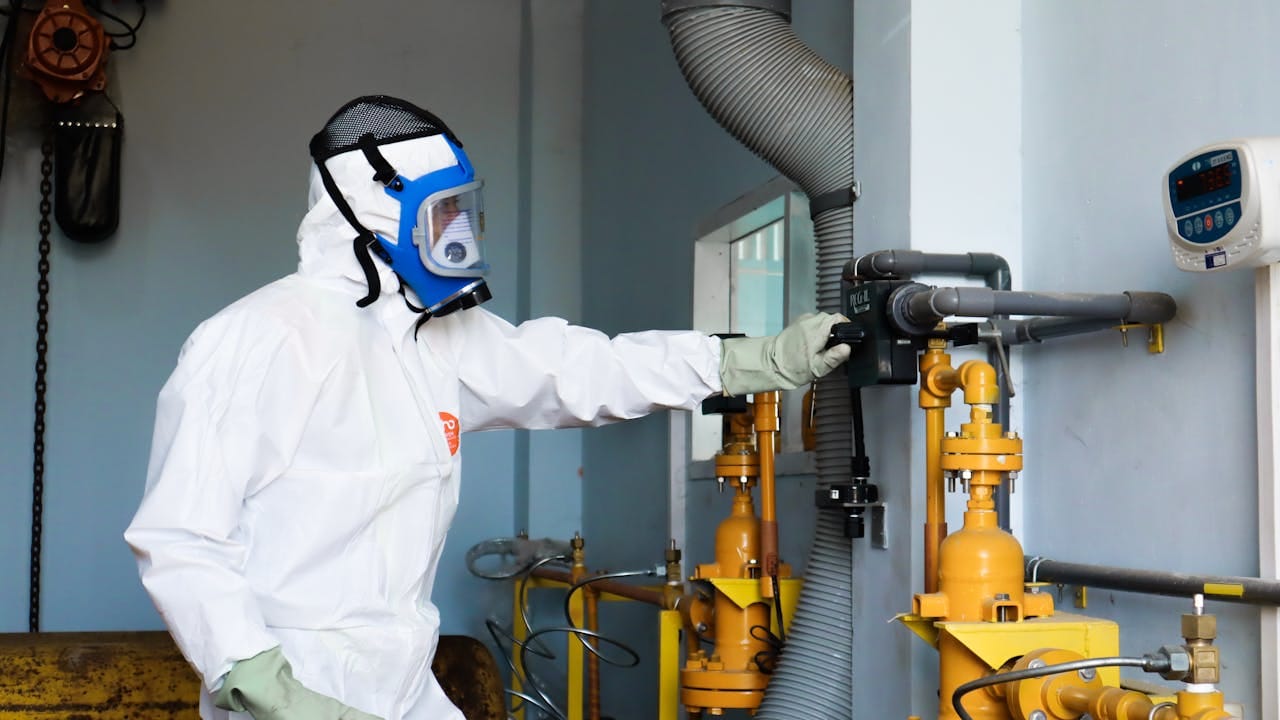 Técnico con traje de protección completo y mascarilla filtrante, operando un sistema de tuberías en un entorno industrial sensible.