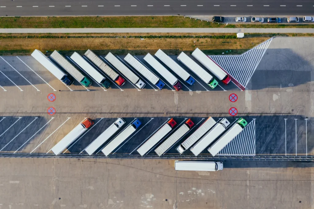 Vista aérea de un aparcamiento para camiones bien organizado, con vehículos pesados estacionados en diagonal en plazas designadas, junto a una carretera principal.