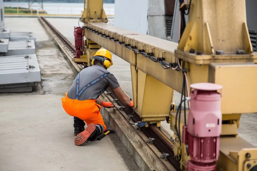 Operario con ropa de alta visibilidad y casco de protección realizando el mantenimiento de una estructura metálica sobre riel en un entorno industrial.