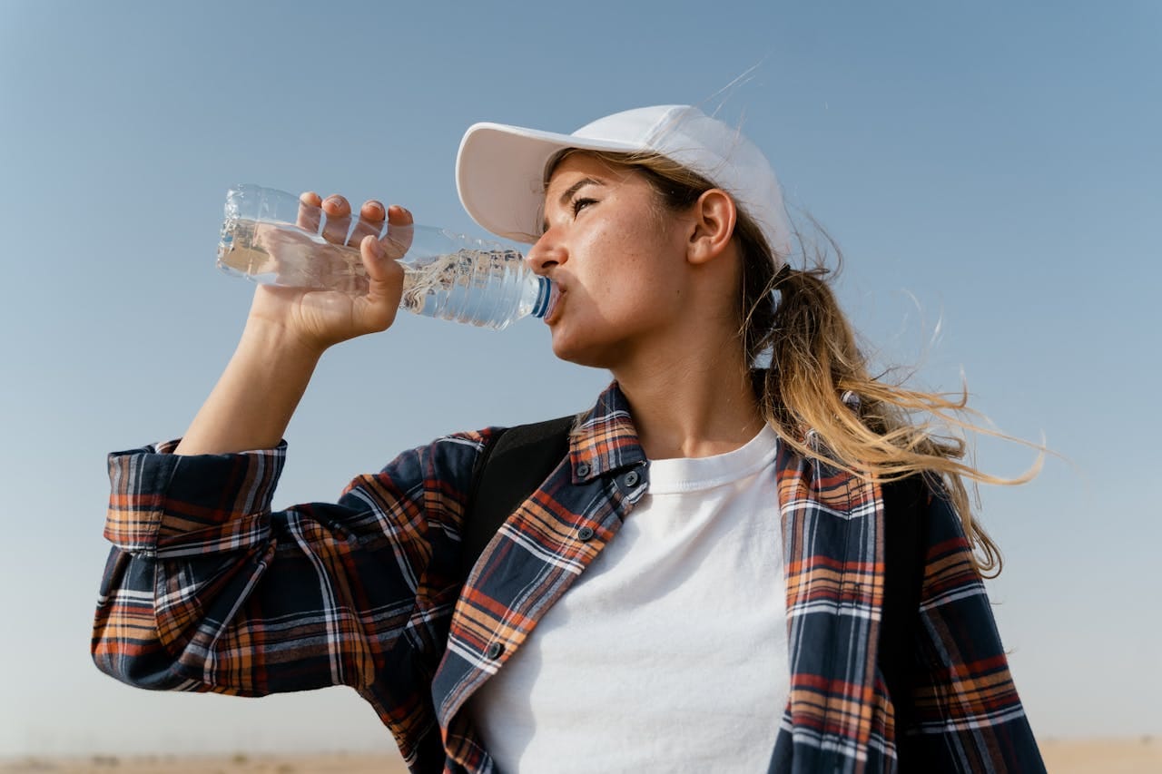 Joven con gorra blanca y camisa de cuadros bebiendo agua bajo el sol para mantenerse hidratada, en un entorno caluroso y seco.