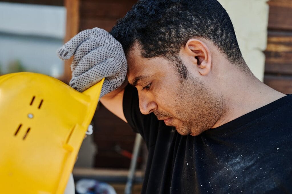 Trabajador de la construcción sudando, con la cabeza agachada y el casco de protección amarillo en la mano, visiblemente afectado por el calor o el esfuerzo físico.